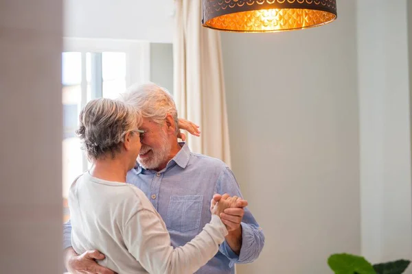 Cheerful senior couple dancing and smiling looking at each other. Elderly happy couple celebrating by dancing in living room. Romantic old aged couple holding hands and dancing together at home