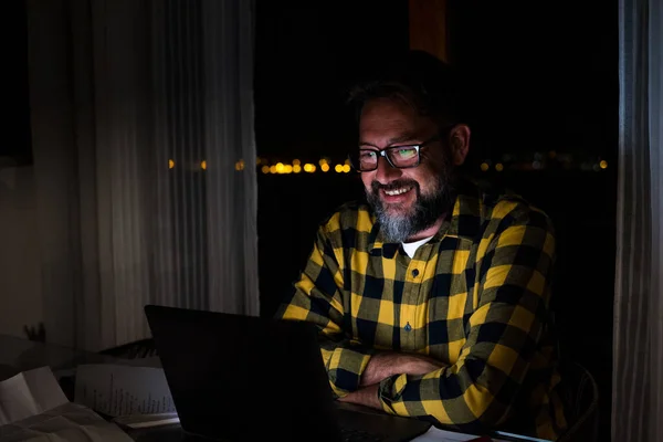 Happy young man using a laptop or computer at home working late night in the dark with the light of the screen on his face smiling