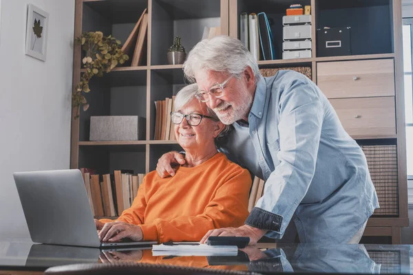 Elderly man and woman having fun using laptop at office. Smiling old husband and wife doing home finance online. Senior couple watching media content and enjoying at home
