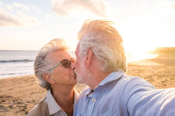 stock image Portrait of couple of mature and old people enjoying summer at the beach looking to the camera taking a selfie together with the sunset at the background. Two active seniors traveling outdoors. 