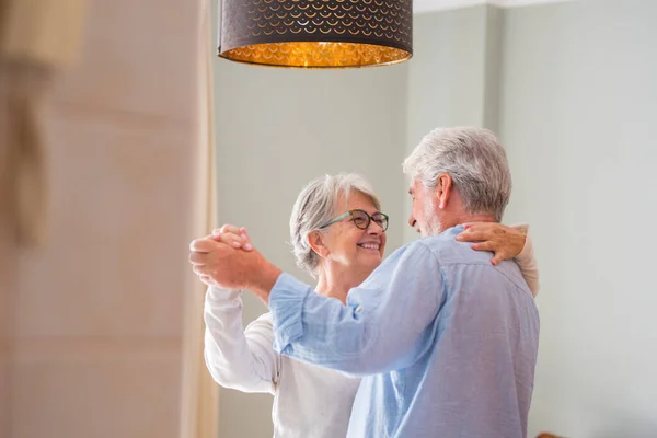 Romantic loving senior couple holding hands enjoying dancing together in the living room of house, Elderly happy couple celebrating by doing dance at home. Old husband and wife having fun time at modern apartment