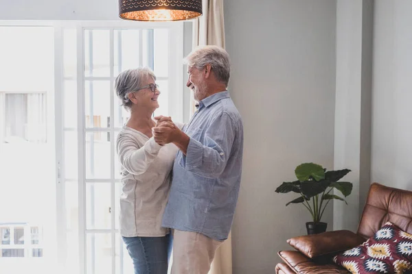 Romantic loving senior couple holding hands enjoying dancing together in the living room of house, Elderly happy couple celebrating by doing dance at home. Old husband and wife having fun time at modern apartment