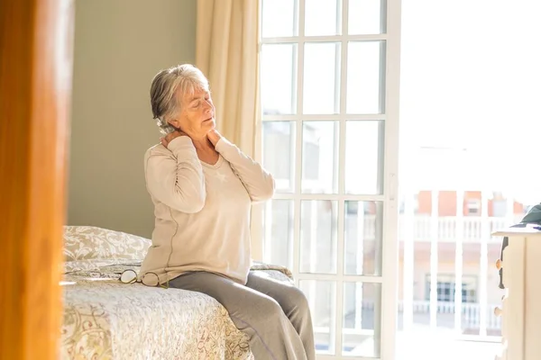 Senior woman suffering from neck ache after sleep, rubbing stiff muscles, Old female sitting on bed touching neck feeling discomfort because of uncomfortable bed at home