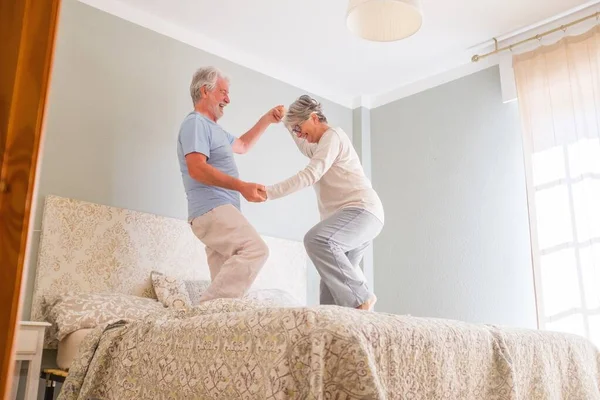 Elderly happy senior couple holding hands and dancing together on bed at home. Carefree active senior heterosexual couple holding hands and dancing together on bed at home