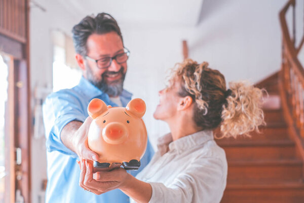 Happy caucasian couple holding piggy bank to save money to make their future dreams come true. Husband and wife holding piggy bank for savings at home while looking at each other 