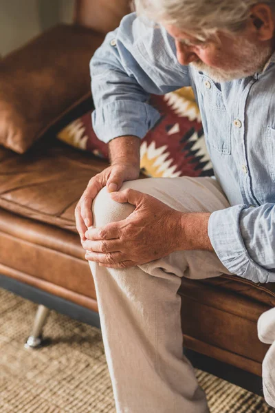 Senior old man facing knee problem, sitting on sofa holding knee at home. Old male suffering from severe joint ache sitting in living room. Healthcare and problem concept