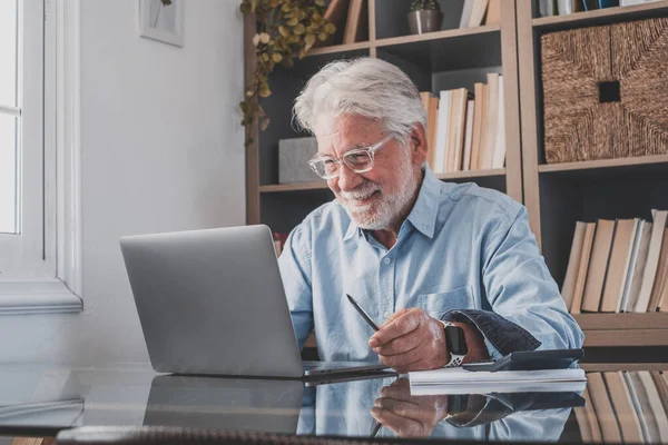 Happy old senior businessman looking at laptop screen and smiling on reading email sitting at home office. Elderly man working online with calculator on table in front of book shelf at workplace