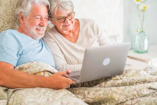 Old senior caucasian couple smiling and video chatting using laptop in the morning at bed in the bedroom at home. Elderly couple greeting on video call on laptop at home.