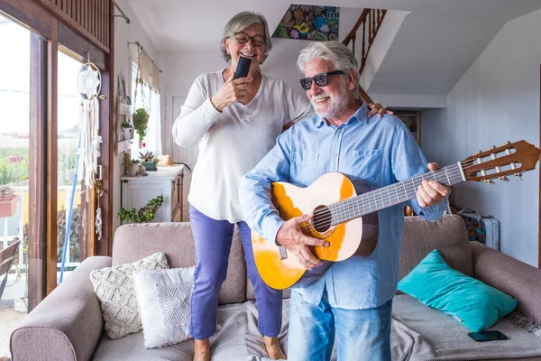 Happy and funny couple of old and mature people having fun and enjoying at home doing a party together singing and dancing playing the guitar indoor. Holiday or even celebrating concept.
