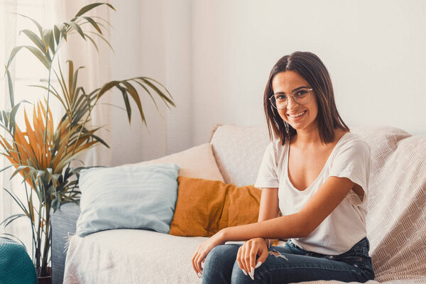 Happy young woman in eyeglasses relaxing on sofa in the living room of her house. Beautiful satisfied lady sitting on couch at modern apartment. Pretty young female looking at camera while sitting at home 