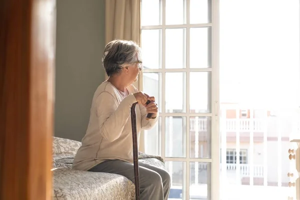 Thoughtful senior woman leaning on walking stick while sitting on bed. Lonely elderly woman sitting in bedroom at home. Old woman with gray hair leaning on walking stick sitting on bed at home