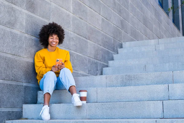 Beautiful african american woman in yellow t-shirt and curly hair using mobile phone outdoors on urban city staircase. Black lady text messaging on smartphone while sitting on steps with disposable coffee cup