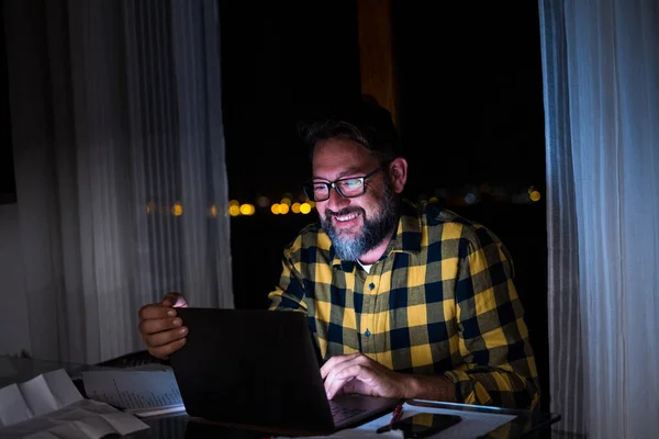 Happy young man using a laptop or computer at home working late night in the dark with the light of the screen on his face smiling