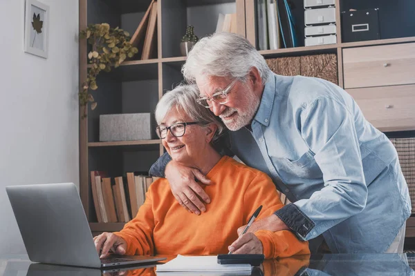 Elderly man and woman having fun using laptop at office. Smiling old husband and wife doing home finance online. Senior couple doing banking work and writing notes at home