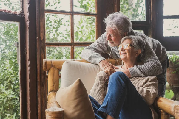 Old caucasian couple spending leisure time looking out through window at home. Loving husband embracing wife from behind sitting on armchair at house and admiring view from transparent door glass.