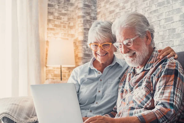 stock image Cute couple of old people sitting on the sofa using laptop together shopping and surfing the net. Two mature people wearing eyeglasses in the living room enjoying technology. Portrait of seniors laughing in love.  