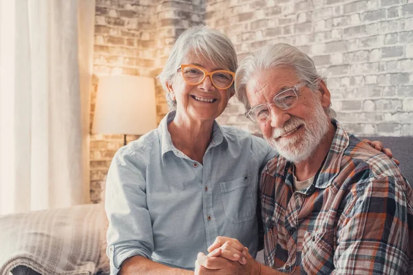 Cute and beautiful couple of old people smiling and looking at the camera having fun at home together. Portrait of seniors wearing eyeglasses sitting on sofa enjoying and relaxing.