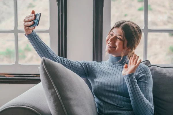 Young beautiful woman sitting on the sofa at home chatting and surfing the net. Female person having fun with smartphone online. Portrait of girl smiling taking a selfie to post it on the social media.