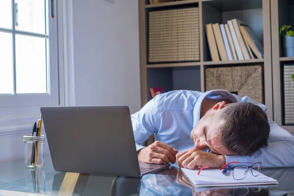 Tired businessman is sleeping at his work desk with laptop computer. Exhausted caucasian guy  lying on table with eyes closed falling asleep. Male executive sleeping at workplace, boring routine work