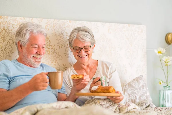 Old senior caucasian couple enjoying breakfast in the morning at bed in the bedroom at home. Elderly couple eating croissant and drinking coffee from cup for breakfast at home.