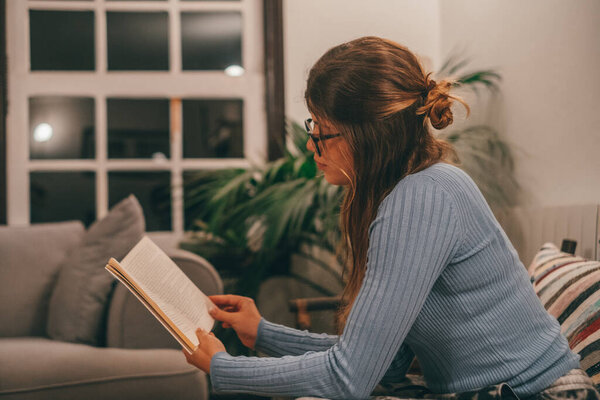 One cute and attractive wearing eye glasses reading and looking an interesting book at home indoor on the sofa. Young woman relaxing alone at night reading books sitting on the couch. 