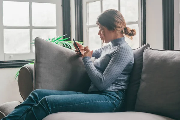 年轻美丽的女人坐在家里的沙发上聊天和上网 在网上玩智能手机的女性 用手机修改社交媒体刻画女孩微笑的形象 — 图库照片