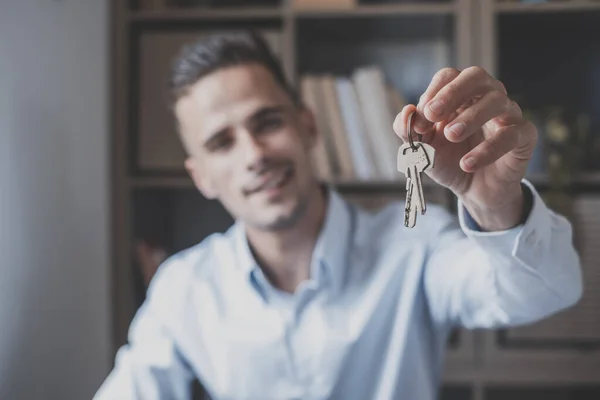 Portrait of smiling young man showing or giving keys of his new house or office. Confident handsome male holding keys of the home or office workplace. Concept of buying or selling a property
