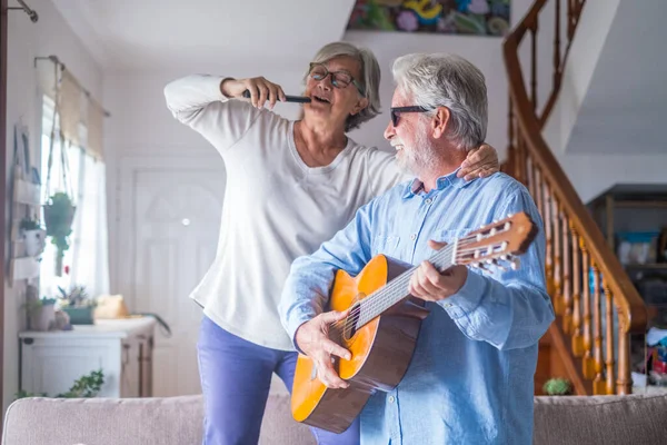 stock image Couple of two happy seniors or mature and old people singing and dancing together at home indoor. Retired man playing the guitar while his wife is singing with a remote control of TV. 