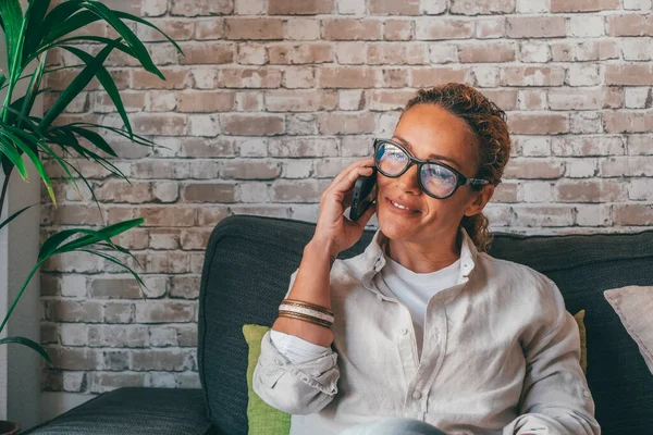 stock image Happy young woman in casual clothes using mobile phone sitting on sofa in the living room of modern luxury apartment. Caucasian lady smiling while talking on smartphone at home 