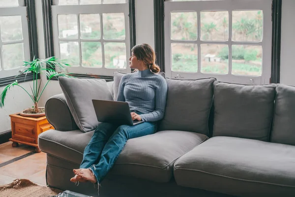One young woman using laptop on the sofa at home looking at the window thinking about future and work. Female thoughtful person relaxing working or surfing the net.