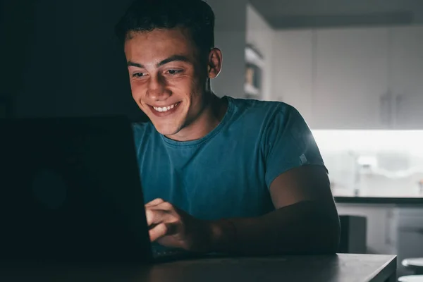 One young teenager studying and working at home at night on the table using laptop pc smiling and having fun doing homework. Light of screen on face.