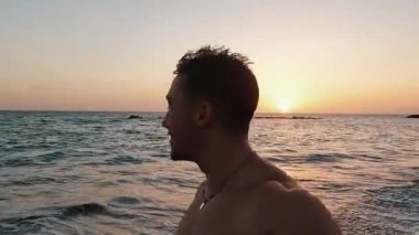 Güzel genç adam kumsalda yürüyor, kameraya bakıp gülümsüyor.