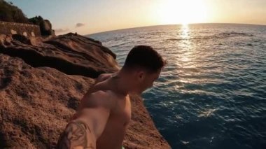 Genç adam uçurumdan atlıyor, kameraya bakıyor ve gülümsüyor. 