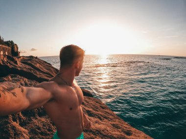 Sahilde tek başına yürüyen neşeli bir genç adam. Arka planda günbatımı var. Erkek insan eğleniyor ve özgürlüğün tadını çıkarıyor. Selfie çekiyor..