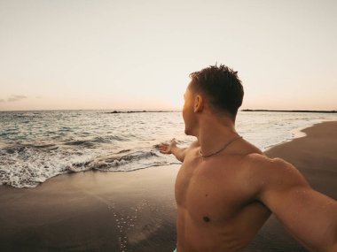 Sahilde tek başına yürüyen neşeli bir genç adam. Arka planda günbatımı var. Erkek insan eğleniyor ve özgürlüğün tadını çıkarıyor. Selfie çekiyor..
