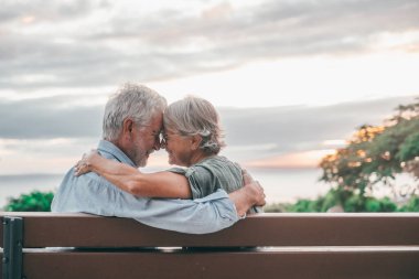 Head shot close up portrait happy grey haired middle aged woman snuggling to smiling older husband, enjoying sitting on bench at park. Bonding loving old family couple embracing, looking sunset.