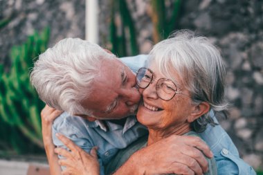 Middle age woman sitting on bench of park with old man hugging her from the back. Couple of retired people relaxing and enjoying sunset together outdoors.