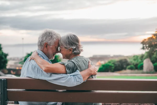 Head shot close up portrait happy grey haired middle aged woman snuggling to smiling older husband, enjoying sitting on bench at park. Bonding loving old family couple embracing, looking sunset.