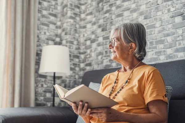 Oturma odasındaki rahat koltukta oturan mutlu, sarışın kadın ilginç kitaplar okuyor. Gülümseyen yaşlı, emekli büyükanne evde yalnız başına en sevdiği romanla hafta sonu tatilinin tadını çıkarıyor..