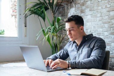 Side view handsome young businessman in eyewear working with computer remotely, sitting at wooden table in office. Pleasant happy man communicating in social network, searching information online.