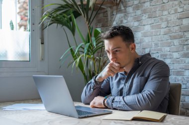 Thoughtful businessman touching chin, pondering ideas or strategy, sitting at wooden work desk with laptop, freelancer working on online project, student preparing for exam at home