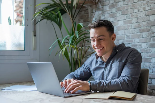 stock image Side view handsome young businessman in eyewear working with computer remotely, sitting at wooden table in office. Pleasant happy man communicating in social network, searching information online.