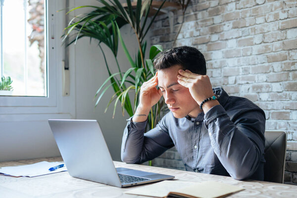 Stressed tired man in pain having strong terrible headache attack after computer laptop work, fatigued exhausted guy suffering from chronic migraine massaging temples to relieve head ache tension
