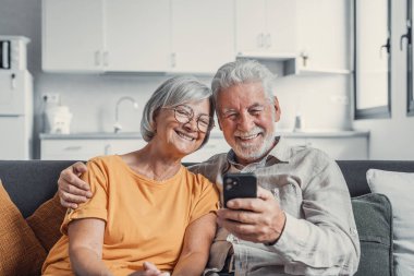 Mutlu olgun aile, eş ve koca birlikte evde telefon kullanarak, kanepede oturan orta yaşlı erkek ve kadın gülümseyen, mobil cihaz uygulamaları kullanarak, sosyal ağda video izlerken, internette sörf