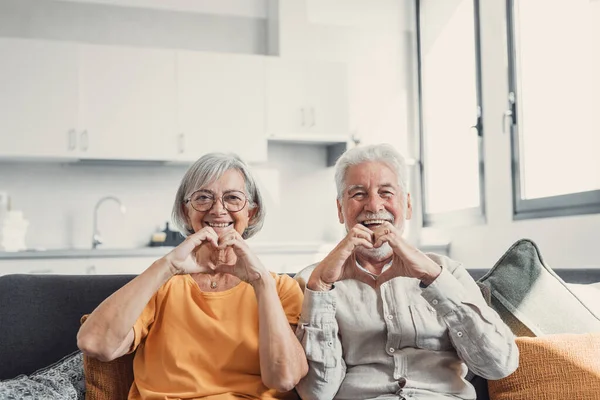 stock image Close up portrait happy sincere middle aged elderly retired family couple making heart gesture with fingers, showing love or demonstrating sincere feelings together indoors, looking at camera.
