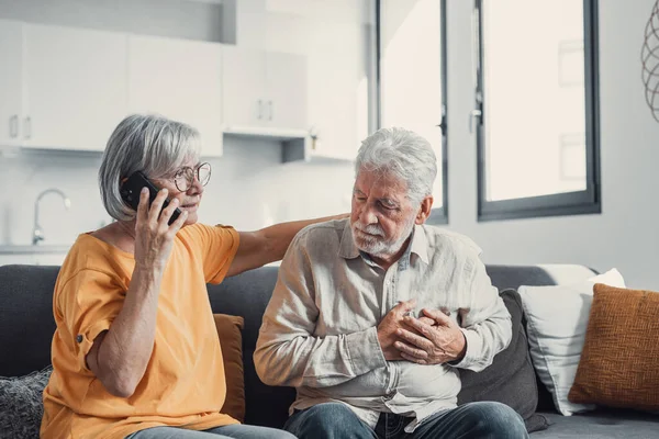 stock image Mature woman, wife calling emergency, talking on phone, holding old husband hand, grey haired man having heart attack, touching chest, suffering from heartache disease at home, feeling pain