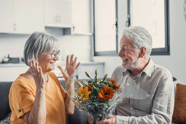Old man giving flowers at his wife sitting on the sofa at home for the San Valentines day. Pensioners enjoying surprise together. In love people having fun