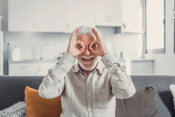 stock image Head shot and portrait of one old mature man sitting on the sofa looking at camera making funny faces with fingers and hands smiling and laughing. Healthy senior enjoying at home