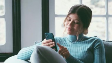 Evdeki kanepede akıllı telefon kullanan güzel bir genç kadının görüntüsü.