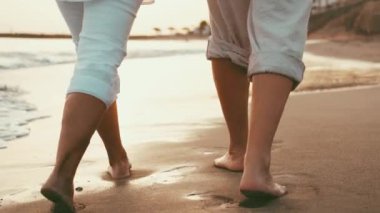 Gün batımında sahilde çıplak ayakla yürüyen romantik bir çiftin kırpılmış görüntüleri.